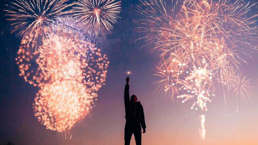 Man with sparkler as fireworks explode behind him